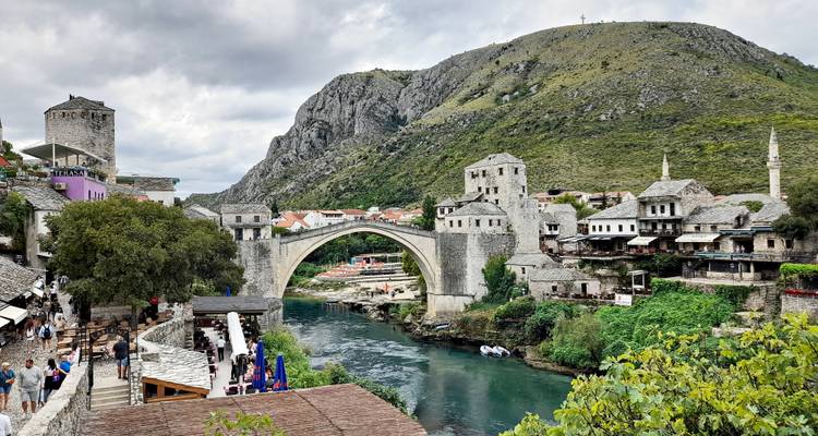 Iconische stenen boogbrug die een turquoise rivier overspant met een oude stad en bergen op de achtergrond.