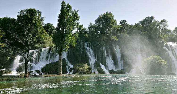 Brede trapsgewijze waterval die naar beneden stort in een mistige poel omringd door weelderig bos en hoge bomen.