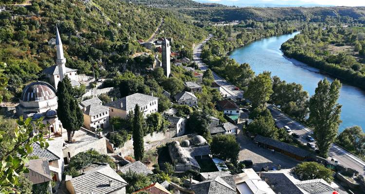 Heuveltopaanzicht over een rivierdorp met minaretten en middeleeuwse stenen torens langs turkoois water.