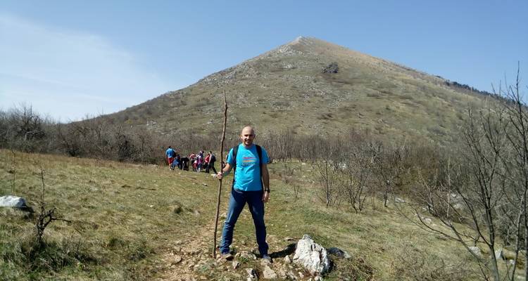 Een man met een wandelstok poseert op een grasrijke berghelling terwijl een rij wandelaars op de achtergrond nadert.