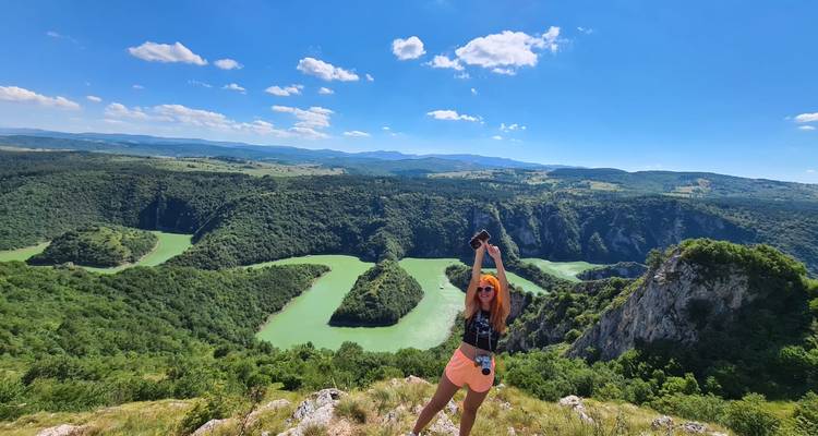 Een glimlachende vrouw steekt triomfantelijk haar armen omhoog op een klif met weids uitzicht op levendige groene rivierbochten en glooiende heuvels.
