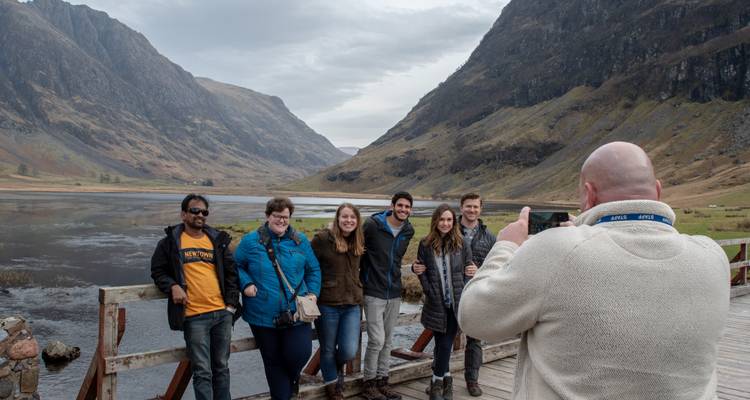 Des amis posent sur un pont en bois devant un loch des Highlands aux reflets miroirs, flanqué de vallées escarpées.