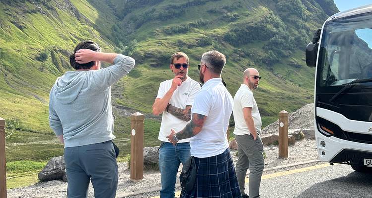 Des voyageurs discutant à côté d'un autocar touristique sur une aire de repos routière au milieu des vallées verdoyantes des Highlands écossaises.