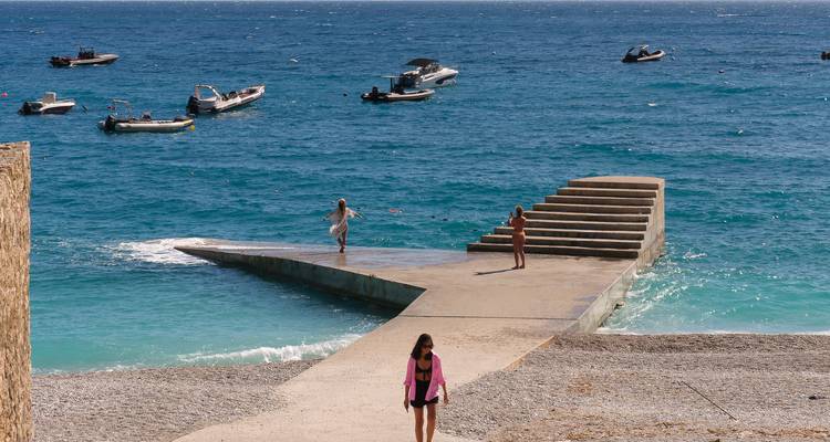 Les gens se promènent et jouent sur une jetée en béton qui s'avance dans une mer d'un bleu éclatant avec de petits bateaux à moteur qui flottent à proximité.