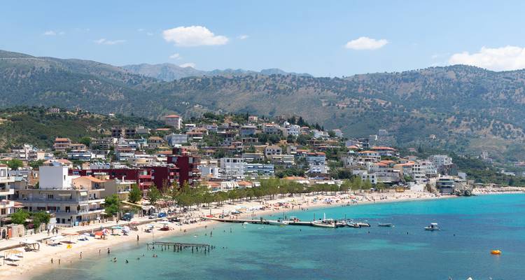 Une ville côtière albanaise avec des montagnes derrière s'étend le long d'une plage animée parsemée de nageurs et de parasols.