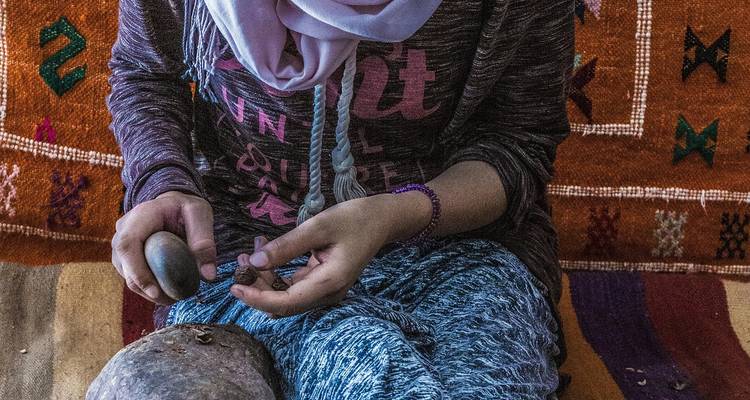 Nahaufnahme einer Frau in traditionellem Kopftuch, die Arganüsse von Hand mit einem Steinwerkzeug mahlt.