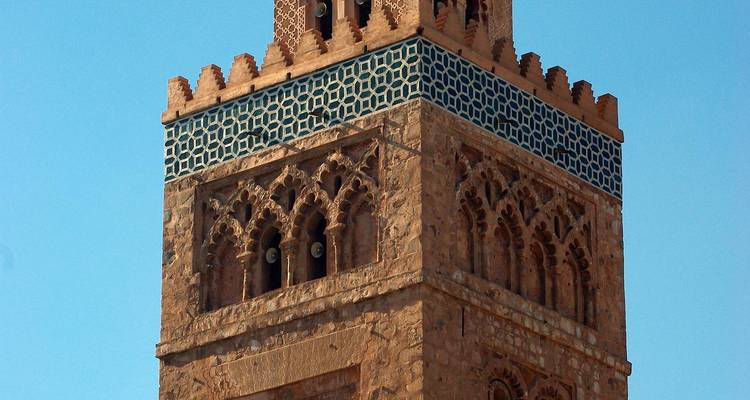 Detailansicht eines historischen Minaretts in Marrakesch mit kunstvollen Schnitzereien und blauen geometrischen Fliesen vor klarem Himmel.
