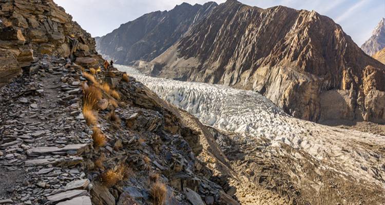Les randonneurs marchent sur un sentier rocheux en altitude au-dessus d'un glacier étendu bordé de montagnes escarpées.
