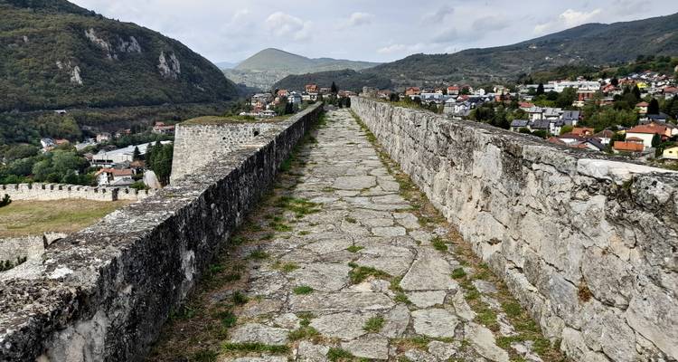 Lange stenen wandelweg langs versterkte muren met uitzicht op een valleistadje en bergen.