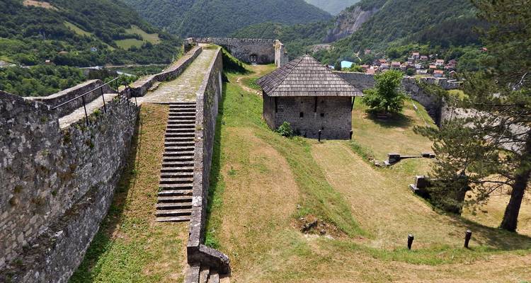 Longs remparts de pierre et escaliers d'une forteresse de colline surplombant des vallées verdoyantes.