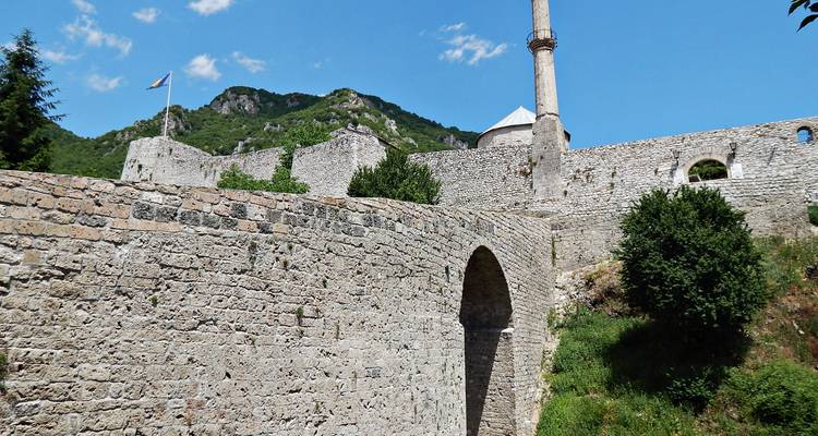 Murs de fort en pierre, pont en arc et un minaret élancé situés sous un ciel bleu de montagne.