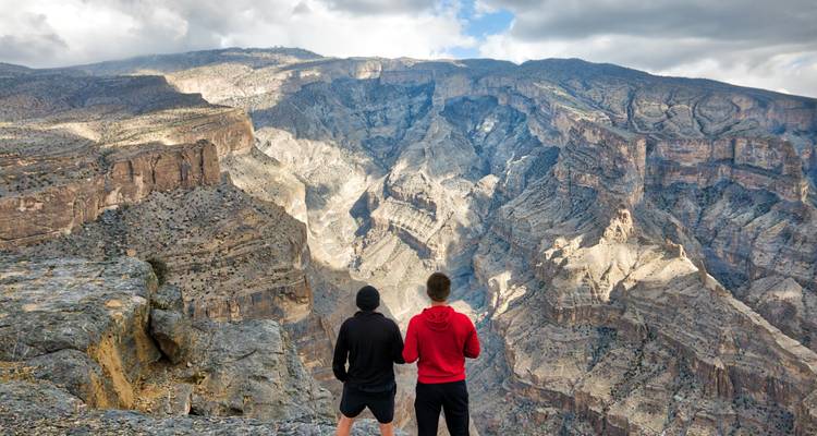 Zwei Wanderer blicken in die weite, zerklüftete Canyonlandschaft des Jebel Shams.