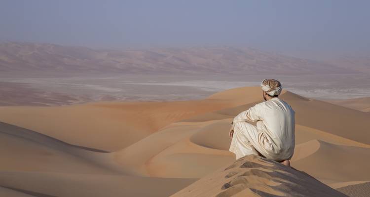 Omanischer Mann in weißem Gewand sitzt nachdenklich auf einer weiten Fläche von sich wellenden Wüstendünen.