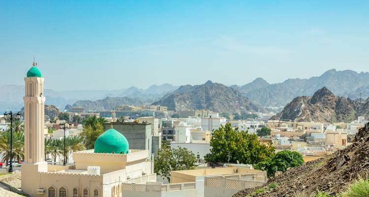 Panoramic view over Muscat with a mosque featuring a green dome and minaret, white low-rise buildings and rugged brown mountains under a clear blue sky.