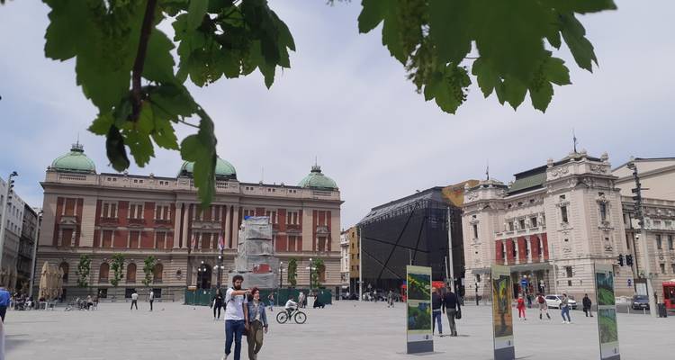 Open stadsplein in Belgrado met historische gebouwen, bladomlijste voorgrond en voetgangers.