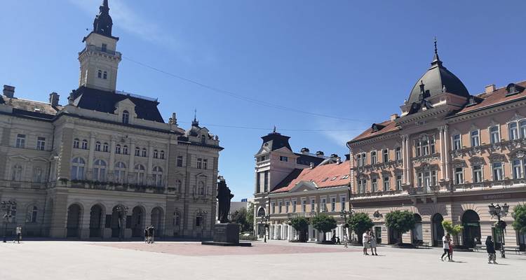 Zonnig hoofdplein van Novi Sad met elegante Oostenrijks-Hongaarse gebouwen en enkele bezoekers.