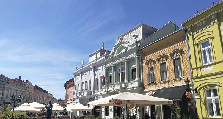 Kleurrijke gevels en terrasjes grenzen aan een charmante voetgangersstraat onder een heldere blauwe hemel in Novi Sad.
