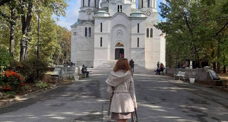 Vrouw in lichte jas loopt naar de witte marmeren Oplenac Kerk omringd door bomen en bezoekers.