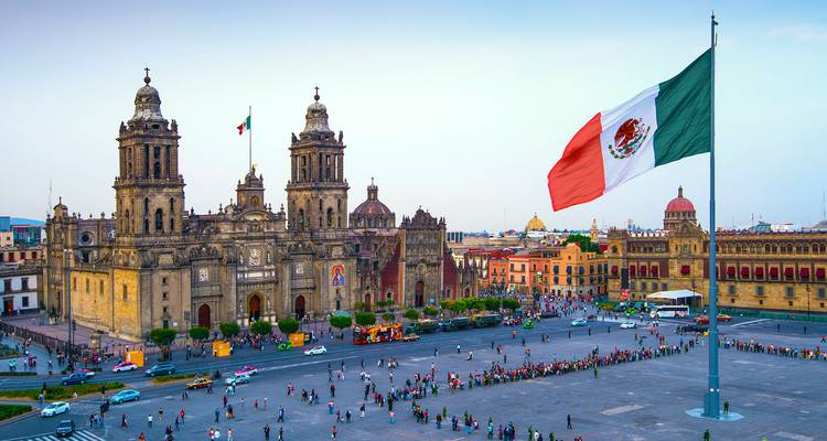 Vue panoramique du Zócalo avec des drapeaux mexicains, des bâtiments historiques et des foules au crépuscule.
