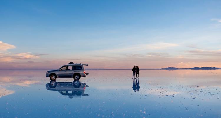 SUV y una pareja se encuentran sobre la superficie espejada del Salar de Uyuni al atardecer, perfectamente reflejados en el salar.