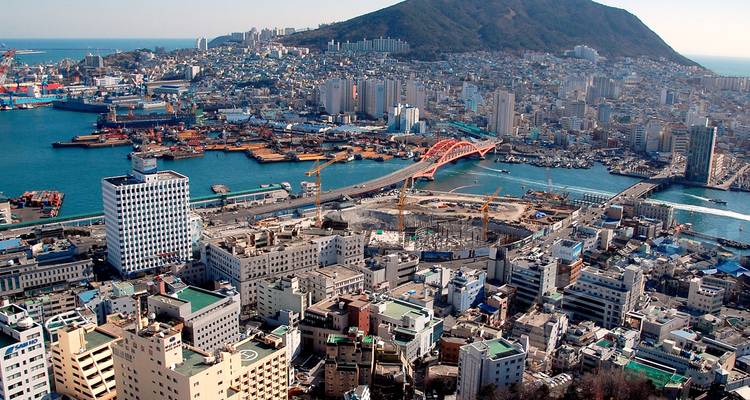 Vue aérienne large d'une ville côtière avec des bâtiments denses, un pont rouge, des quais portuaires animés et une montagne boisée s'élevant derrière l'horizon par une journée claire.