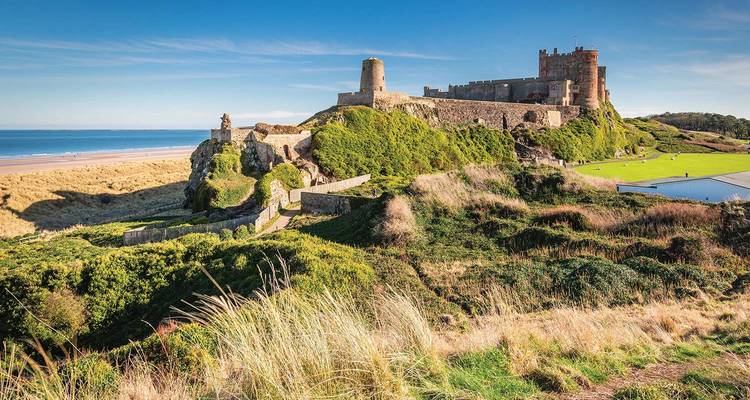 El castillo medieval de Bamburgh encaramado en un acantilado cubierto de hierba con vista a una playa de arena y el mar azul.