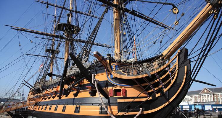 Buque de guerra histórico de madera con mástiles imponentes y casco negro y naranja atracado en un puerto naval.