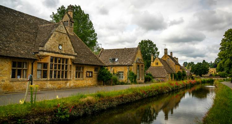 Pintorescas casas de campo de piedra color miel reflejadas en un canal estrecho bajo un cielo nublado de los Cotswolds.