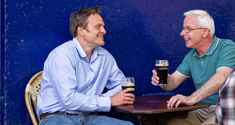 Dos hombres compartiendo pintas de Guinness en una mesa de pub al aire libre contra una pared azul profundo.