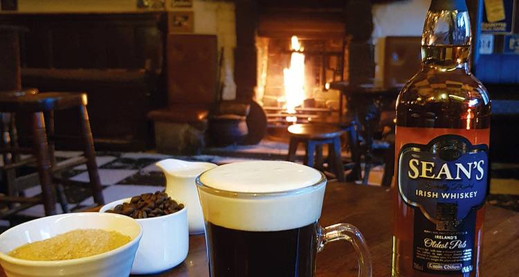 Acogedora escena de pub irlandés con una copa de café irlandés, botella de whisky y fuego abierto al fondo.