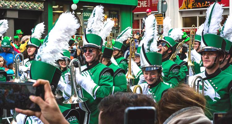 Banda de música colorida vestida con uniformes verdes y sombreros con plumas blancas actuando durante un desfile urbano animado.