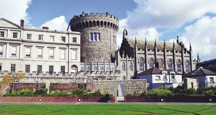 Torre redonda de piedra histórica y capilla gótica del Castillo de Dublín bajo un cielo azul con césped cuidado en primer plano.