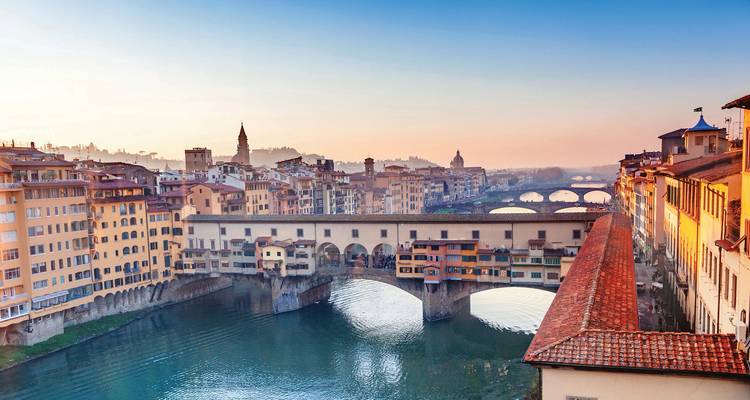 Puente histórico de piedra bordeado de edificios que cruza un río tranquilo al amanecer