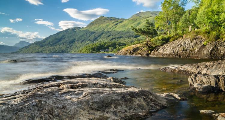 Les vagues claires du lac lèchent le rivage rocheux sous les montagnes écossaises boisées par une journée lumineuse