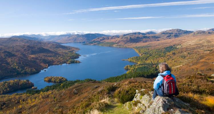 Randonneur avec sac à dos rouge assis sur un affleurement rocheux surplombant un vaste loch bleu et un paysage de hautes terres vallonnées