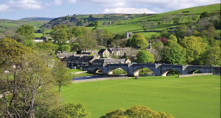 Un pont de pierre enjambe une rivière sinueuse à côté d'un pittoresque village anglais niché dans des vallons verdoyants.