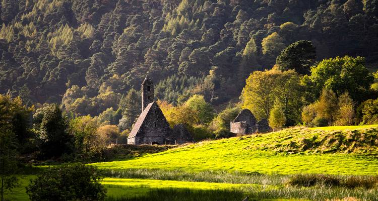 Stone monastic church amidst vibrant green meadow with forested hills lit by warm sunlight