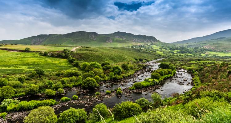 Wide green valley with a winding river and distant rolling hills under a cloudy sky.