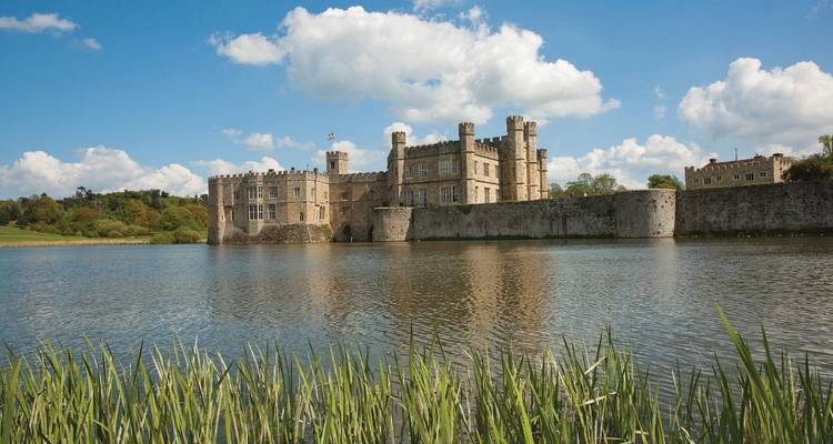 Château de Leeds reflété dans des douves immobiles encadrées de roseaux sous un ciel lumineux.