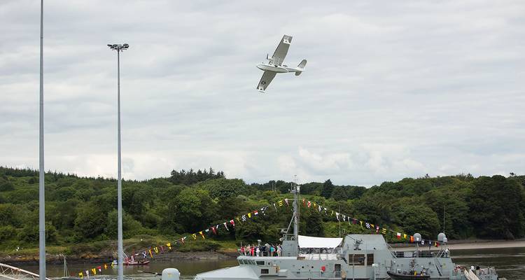 Un petit aéronef effectue une manœuvre de spectacle aérien au-dessus d'un navire militaire amarré le long d'un rivage boisé.