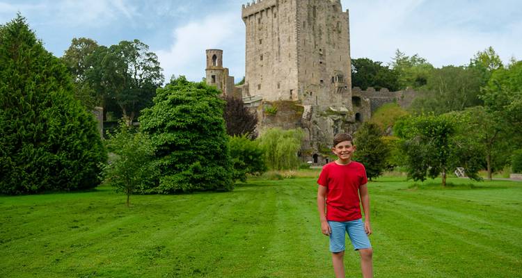 Smiling boy stands on green lawn with imposing Blarney Castle looming behind.