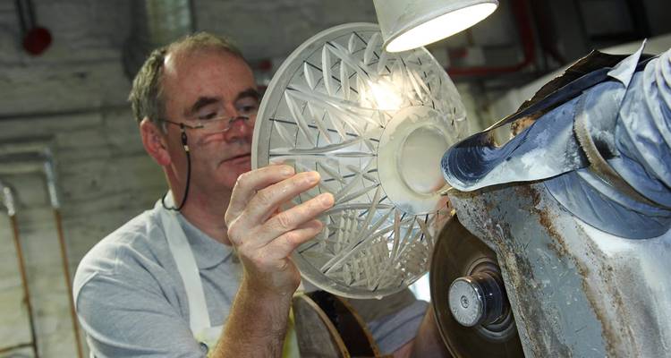 Craftsman carefully grinds intricate patterns into a crystal bowl under a workshop lamp.