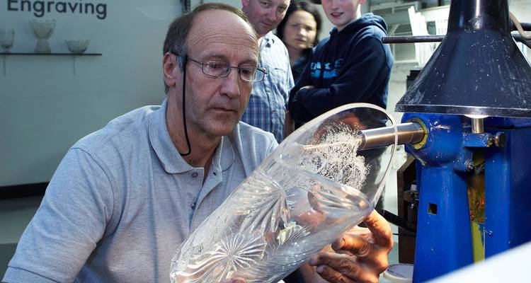 Glass engraving specialist demonstrates carving on a crystal vase while onlookers observe closely.