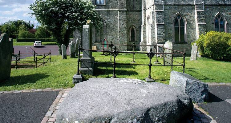Large glacial boulder and iron railing outside stone church building in green churchyard.