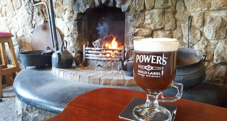 Irish coffee with creamy head sits on a wooden table before a lit stone fireplace in a cozy pub.