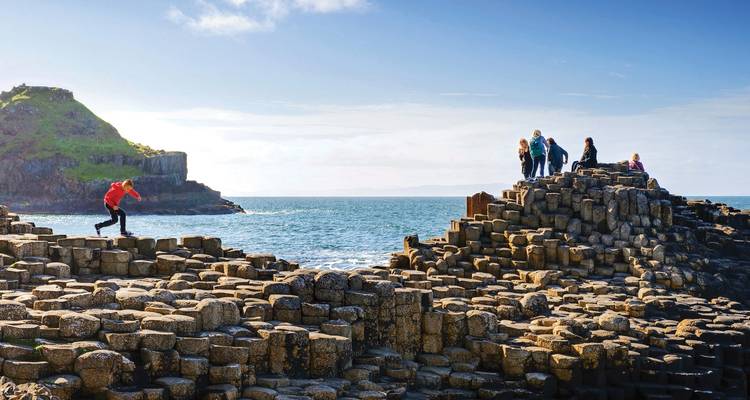Travelers scale basalt columns of the Giant's Causeway under bright blue skies and sunshine.