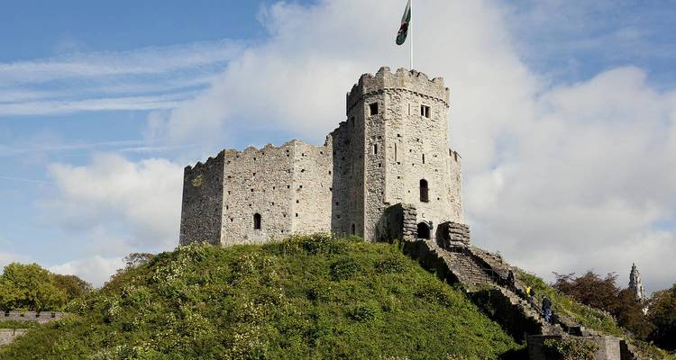 Donjon de château de pierre médiéval perché sur une butte herbeuse sous un ciel bleu éclatant, avec un drapeau gallois flottant au sommet.
