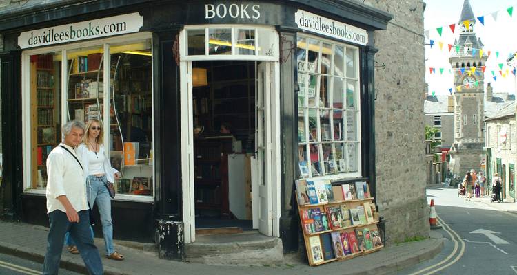 Charmante librairie d'angle avec de grandes vitrines exposant des livres, des banderoles colorées au-dessus et deux jeunes visiteurs qui passent devant.