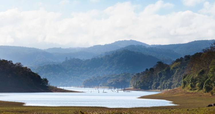 Lac serein serpentant entre des collines boisées et des montagnes lointaines sous de doux nuages.