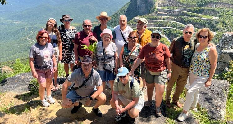 Un grupo de turistas sonrientes posa en un mirador rocoso sobre una dramática carretera serpenteante de montaña.